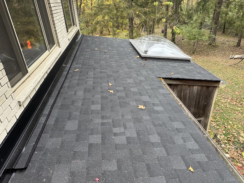 Modern flat roof with dark gray shingles and skylight in a vibrant autumn landscape.